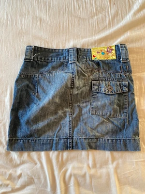 Blå jeanskjol från Vingino - Snygg blå jeanskjol från Vingino med coola fickor fram och bak, bälteshällor och en blomformad knapp. Kjolen har broderad etikett med fjärilar och Vingino-logga. Perfekt för dig som gillar denim och detaljer. Vintage, storlek 30 i midja men liten 