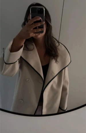 Beige kappa med svarta detaljer - Säljer en beige kappa med bred krage och svarta kantdetaljer. Helt oanvänd. Från Bubbleroom