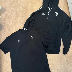 Svart Adidas Juventus hoodie - Svart hoodie från Adidas med Juventus-logga och vita ränder längs ärmarna. Dragkedja framtill, snörning i huvan och två fickor. Adidas-logga på bröstet. Perfekt för dig som gillar sportig stil och fotboll.