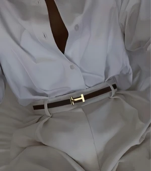 Brunt skärp med H-spänne - Säljer ett stilrent brunt bälte i skinn med ett ikoniskt guldigt H-spänne. Bältet är smalt och har snygga sömmar längs kanterna. Perfekt för att lyfta din outfit med en lyxig detalj och klassisk design.