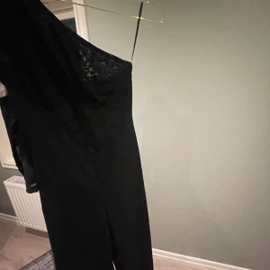 Svart långklänning med oneshoulder - Säljer en svart byxdress med oneshoulder-design och lång ärm. Klänningen har en stilren look med lätt transparent tyg på axeln och en figurnära passform. Perfekt för dig som vill sticka ut med en elegant och modern stil.