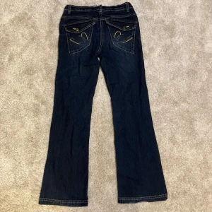 Mörkblå bootcut jeans från Here+There - Snygga mörkblå jeans från Here+There med bootcut passform och coola gula broderier. Perfekt för dig som gillar en lite retro vibe och vill sticka ut med detaljer. Lite slitet vid ena benet längst ner men det är inget som märks