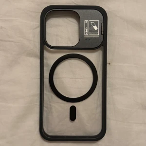  iPhone 15 Pro - Transparent MagSafe-kompatibelt skal med svart ram och inbyggt kameraskydd. Passar iPhone-modeller med stor kameramodul. Skalet är i nyskick utan synligt slitage, och kameraskyddet är lätt att öppna och stänga för extra skydd.