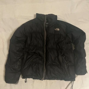 Svart pufferjacka från The North Face - Säljer en svart pufferjacka från The North Face, modell 550. Jackan har hög krage, dragkedja framtill och två sidofickor. Den är vadderad och har justerbar nederkant med snörning. Perfekt för kalla dagar och har en clean, sportig look. Passar som M