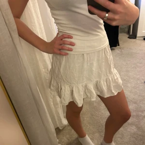 Vit kortkjol med volangkant - Supersöt vit kortkjol med volangkant längst ner. Kjolen har en lätt skrynklig struktur och är gjord i ett mjukt bomullsmaterial som känns skönt mot huden. Perfekt för dig som gillar en feminin och chill stil. Elastisk midja för skön passform.