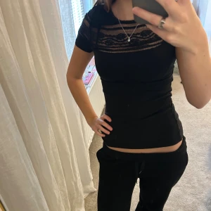 Svart topp med spetsdetalj - Säljer en svart topp med korta ärmar och snygg spets upptill över bröstet. Modellen är tight och har rund halsringning. Spetsen har ett diskret mönster som ger en cool touch. Perfekt att styla med jeans eller kjol för en trendig look.