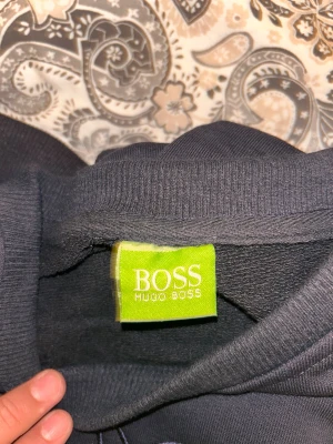 Svart sweatshirt från Hugo Boss - Svart sweatshirt från Hugo Boss med stor broderad logga i mörkblått och neongrönt på bröstet. Tröjan har rund hals och ribbade muddar. Materialet känns mjukt och är troligtvis bomull eller polyester. Perfekt för dig som gillar stilrena och snygga märkesplagg. Står att den är storlek XXL men sitter som L