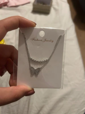 Silverfärgat halsband med fjäril - Snyggt silverfärgat halsband med två kedjor, där den ena har en rad små stenar och den andra en glittrig fjäril som hänge. Perfekt för dig som gillar detaljer och vill ha något som sticker ut lite extra.