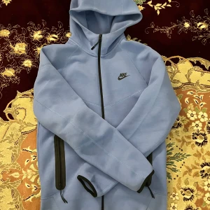 Ljusblå Nike hoodiejacka S - Nike hoodiejacka i ljusblått med svart dragkedja och logga på bröstet. Jackan har huva, långa ärmar och praktiska fickor med dragkedja. Tillverkad i mjuk bomullsmix, perfekt för chill dagar eller när du är på språng.