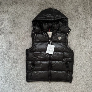 Svart dunväst från Moncler med huva - Svart dunväst från Moncler med avtagbar huva och klassisk logga på bröstet. Västen har quiltad design, två dragkedjeförsedda fickor och hel dragkedja framtill. Materialet är glansigt.