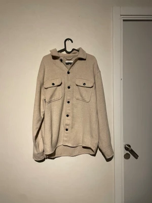 Beige overshirt med svarta knappar - Snygg beige overshirt med svarta knappar och två stora bröstfickor. Oversized passform och mjukt material som känns skönt mot huden. Perfekt att slänga på sig över en t-shirt. Långärmad och med klassisk krage för chill stil.