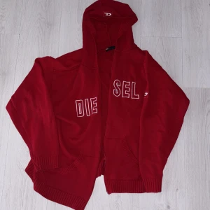 Röd hoodie från Diesel med huva - Säljer en snygg röd hoodie från Diesel med stor huva och broderad logga i vitt på framsidan. Tröjan är stickad och har ribbade muddar samt fickor framtill. Perfekt för dig som gillar streetwear och vill ha något som sticker ut, dragkedjan saknas.nypris 1399