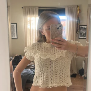 Virkad blus med volang  - Superfin vit stickad blus från Zara i storlek S. Blusen har korta ärmar, högt ribbat halslinne och en bred volangdetalj över axlarna och bröstet. Materialet är stickad bomull med ett snyggt hålmönster och volangen är i broderad spets. Perfekt för dig som gillar romantisk stil!