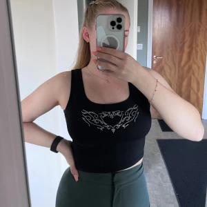 Svart croppad linnetopp - Svart croppad linnetopp med breda axelband och ett  hjärtmönster i silverfärgade strass på bröstet. Toppen har en tight passform och är tillverkad i mjuk bomull med lite stretch. Perfekt för dig som gillar edgy detaljer och vill sticka ut.