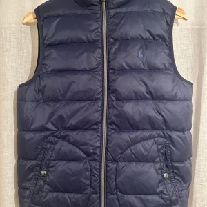 Blå och grå dunväst från Polo Ralph Lauren - Snygg och quiltad dunväst från Polo Ralph Lauren som är vändbar med en marinblå och en ljusgrå sida. Västen har dragkedja framtill, två fickor med knappstängning och den klassiska broderade Polo-loggan på bröstet. Perfekt för lager-på-lager och chill stil. Den är i storlek L (14-16). Den har ett litet hål