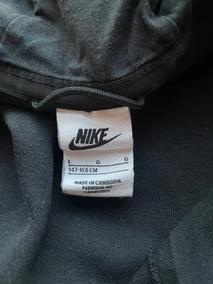Svart Nike tech fleece med dragkedja - Svart hoodie från Nike med hel dragkedja och huva. Snygga gråa ränder över bröstet och ärmen, samt diskret Nike-logga på vänster sida. Jackan har lång ärm och är gjord i mjuk bomull, perfekt för chill eller träning.