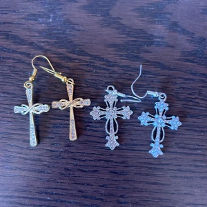 Korsörhängen i guld och silver - Två par snygga örhängen med korsdesign, ett par i glittrande guld med strassdetaljer och ett par i silver med dekorativa stenar. Perfekt för dig som gillar statement-smycken och vill sticka ut med coola detaljer. Säljes antingen enskilt för 30 kr styck eller 50 kr för båda!