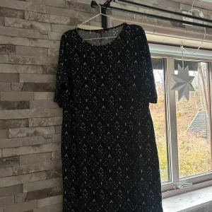 Svart mönstrad klänning Indiska XL - Svart klänning från Indiska i storlek XL med vitt dekorativt mönster. Modellen är rak och har korta ärmar samt rund halsringning. Materialet känns mjukt och stretchigt, troligen bomull eller polyester. Perfekt för dig som gillar grafiska prints och enkel stil.
