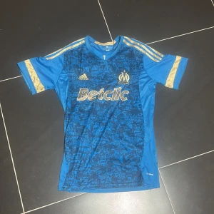 Blå Marseille fotbollströja Adidas 2014 modell - Säljer en blå Olympique de Marseille fotbollströja från Adidas med guldiga detaljer och korta ärmar. Framsidan har ett mörkblått mönster, den är använd 2 gånger. Så gott som ny