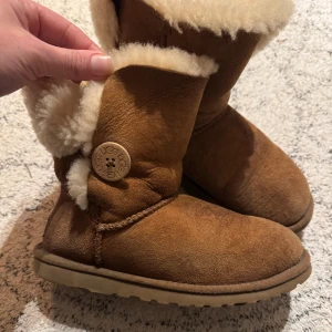 Beiga UGG boots med fårskinn - Mysiga bruna UGG boots med fluffigt vitt fårskinnsfoder och dekorativ knapp på sidan. Gjorda i mjuk mocka med platt sula och rund tå. Perfekta för kalla dagar när du vill hålla fötterna varma och sköna.