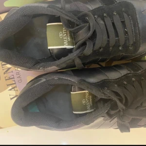 Valentino Garavani svarta sneakers - Svarta Valentino Garavani sneakers med snygga camouflagemönstrade detaljer. Skorna har snörning, platt sula och rund tå. Materialmix av skinn och mocka ger en lyxig känsla. Perfekt för dig som vill ha en cool och exklusiv look och perfekt nu inför vintern med en högre sko för att undvika få in snö och vatten i skon. Box ingår.