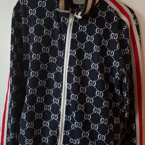 Gucci dress - Gucci dress mörkblått med helvitt GG-mönster och röda samt vita ränder längs ärmarna. Jackan har dragkedja framtill, ribbade muddar och krage med marinblå och vita ränder. Materialet är mjukt och känns lyxigt, perfekt för dig som vill sticka ut. Storlek XL men passar L också. Inte äkta.