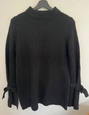 Svart stickad tröja - Svart stickad tröja från Rut&Circle i storlek S. Tröjan har grov ribbad struktur, långärmade ärmar och snygga rosetter vid ärmsluten. Hög krage som ger extra mysfaktor och stil. Perfekt för kyliga dagar och enkel att matcha med jeans eller kjol.