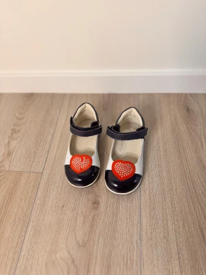 Choupette ballerinaskor med hjärta - Söta ballerinaskor från Choupette i vitt och mörkblått skinn med kardborreband. Skorna har rund tå och är dekorerade med ett rött hjärta med glittriga stenar framtill. 