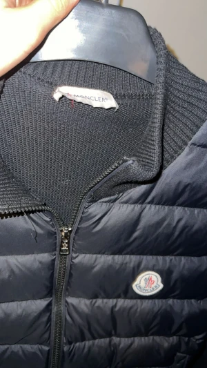 Svart Moncler cardigan jacka med dun - Svart cardigan jacka från Moncler med dunfylld front och stickade ärmar och rygg. Jackan har dragkedja framtill, ribbad krage och Moncler-logga på bröstet. Perfekt för dig som vill ha en stilren och exklusiv look.
