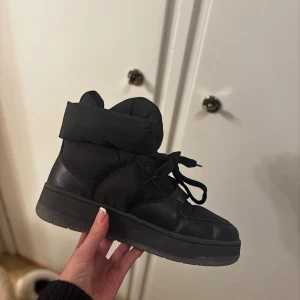 Svarta höga sneakers med snörning - Svarta sneakers med högt skaft och snörning. Skorna har en vadderad, puffig krage och platt sula. Materialet är en mix av skinn och syntet, vilket ger en cool och chunky look