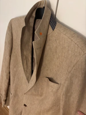 Beige linnékavaj från Morris - Snygg beige kavaj från Morris i linne med klassisk passform. , fyra knappar vid ärmslut . Kragen har ett mönstrat tyg på insidan som ger en extra touch. Perfekt för dig som vill ha en  riktigt go linne kavaj. Nypris cirka 2500 