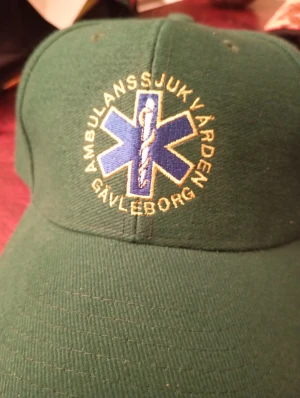 Keps Ambulanssjukvården Gävleborg - Snygg grön keps med broderad blå stjärna och gul text 'Ambulanssjukvården Gävleborg' framtill. Klassisk böjd skärm och ventilerande hål upptill. Materialet känns som bomull och kepsen har en normal passform. Perfekt för dig som gillar unik och officiell merch.