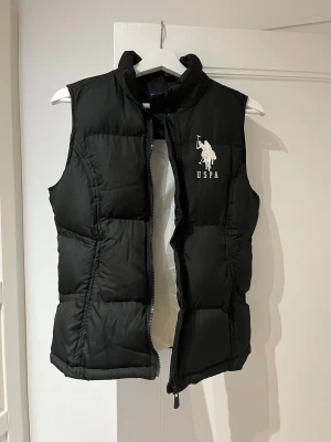 Svart dunväst från U.S. Polo Assn. - Svart dunväst från U.S. Polo Assn. med vit broderad logga och polospelare på bröstet. Västen har hög krage, quiltad design och dragkedja framtill. Tillverkad i 100% polyester, perfekt för lager-på-lager under kyliga dagar.