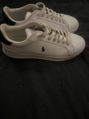 Vita sneakers från Polo Ralph Lauren - Snygga vita sneakers från Polo Ralph Lauren med svart detalj på hälen och klassisk logga på sidan. Skorna har snörning, rund tå och platt sula. Tillverkade i skinn för en clean och stilren look. Perfekta för dig som gillar enkel och tidlös stil.