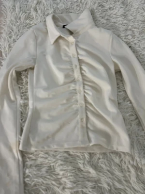 Vit skjorta med rynkade detaljer BikBok - Snygg vit skjorta från BikBok i storlek XS. Skjortan har långa ärmar, klassisk krage och knappar framtill. Materialet är mjukt och har en lätt glans. Rynkade detaljer längs knappraden ger en cool och modern look.