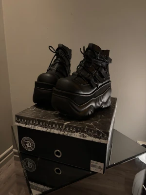 Svarta plattform boots från Demonia - Säljer ett par svarta boots från Demonia med rejäl plattformssula och chunky design. Skorna har snörning, spännen och är tillverkade i syntetmaterial med matt finish. Perfekt för dig som gillar edgy och alternativ stil.