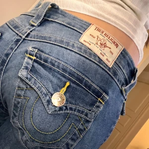 True Religion Joey Low Rise Flare Jeans - Säljer ett par blå True Religion Joey jeans som är bootcut . Är för övrigt i bra skick förutom att det har blivit hål i skrevet. Dessa hål har blivit lagade i butiken vi köpte jeansen ifrån men man kan fortfarande se att det har varit hål där förut som blivit lagade. Man ser inte de lagade hålen när man har på sig jeansen! 