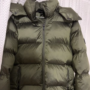 Olivgrön pufferjacka från Everest - Säljer en olivgrön pufferjacka från Everest med stor huva, dragkedja framtill och två sidofickor med dragkedja. Jackan har ett snyggt badge på ärmen och är tillverkad i glansig polyester. Perfekt för kalla dagar med sin tjocka och fluffiga look.
