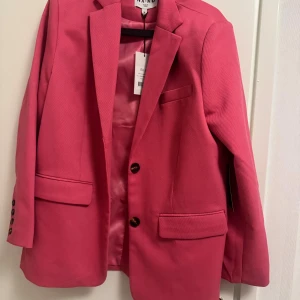 Oversized rosa kavaj från NA-KD - Snygg oversized kavaj från NA-KD i stark rosa färg. Kavajen har klassisk krage, två framfickor med lock, bröstficka och knappar i mörk ton på ärmslut och framtill. Materialet är twill och insidan är fodrad i matchande rosa. Perfekt för dig som vill sticka ut.