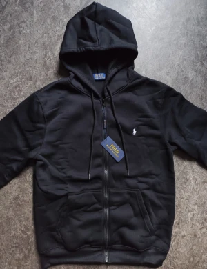 Svart hoodie från Polo Ralph Lauren - Svart hoodie från Polo Ralph Lauren med klassisk huva och snörning, dragkedja framtill och två fickor. Liten vit logobrodering på bröstet. Tillverkad i mjuk bomullsmix, perfekt för chill dagar. Snygg och enkel design som funkar till allt.