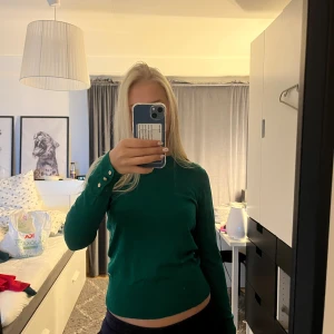 Grön stickad tröja Holly & Whyte - Snygg grön stickad tröja från Holly & Whyte med rund hals och långa ärmar. Tröjan har tre guldfärgade knappar på mudden vid ärmslutet som cool detalj. Passformen är normal och materialet är mjuk bomull. Perfekt för dig som gillar stilrena och enkla plagg.