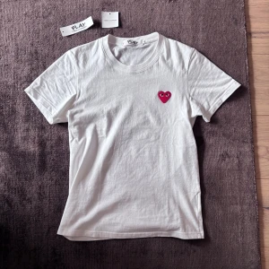 Vit t-shirt Comme des Garçons Play S - Säljer en vit t-shirt från Comme des Garçons Play i storlek S. T-shirten har ett rött hjärta med ögon broderat på bröstet och är tillverkad i mjuk bomull. Klassisk rund hals och korta ärmar, perfekt för en clean och stilren look.