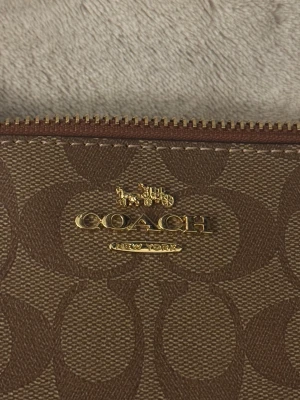 Brun axelväska från Coach - Snygg brun axelväska från Coach med klassiskt monogrammönster och guldfärgade detaljer. Väskan har en smidig dragkedja upptill och ett brunt läderhandtag med Coach-tag. Perfekt storlek för det viktigaste och stilren look.