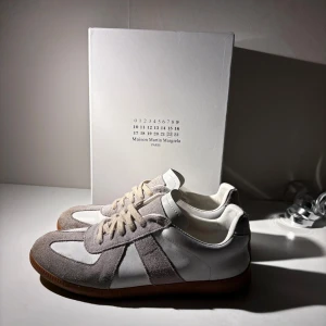 Maison Margiela skor  - Ett par väldigt snygga och trendiga Maison Margiela skor i färgen vit och beige/grå. Dom är i ett bra skick trots lite användning. Det är dock ett litet hål i sulan som kommer från att jag använt dom. Box och dustbag följer med såklart!