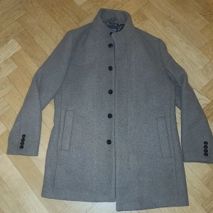 Grå rock från Dressmann, storlek 54 - Snygg grå rock från Dressmann med rak passform och ståkrage. Jackan har svarta knappar framtill och på ärmsluten, samt två diskreta sidofickor. Tillverkad i en mjuk ullblandning som ger en klassisk och stilren look. https://dressmann.com/sv/p/ytterplagg-kappor--rockar-herr-collin-ullrock-mockabrun/7364323_F861   Nypris är ca 2500kr