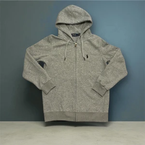 Grå hoodie från Polo Ralph Lauren - Snygg grå hoodie från Polo Ralph Lauren med klassisk svart logga broderad på bröstet. Tröjan har huva med snörning, dragkedja framtill och ribbade muddar vid ärmar och nederkant. Tillverkad i mjuk bomull för skön komfort och stil. M men passar S.