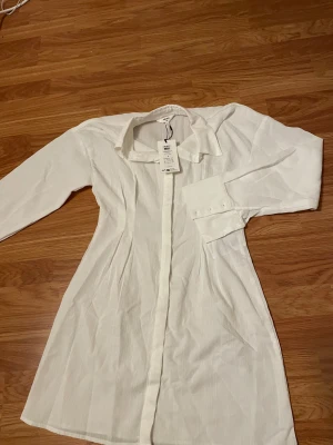 Vit långärmad skjortklänning Object - Snygg vit skjortklänning från Object i storlek 38. Modellen har klassisk krage, långa ärmar med breda manschetter och knappar framtill. Materialet är lätt och känns fräscht, perfekt för dig som gillar stilrena och enkla plagg.