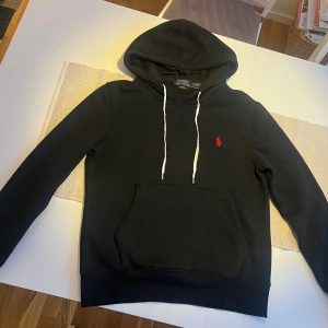 Svart hoodie från Polo Ralph Lauren - Svart hoodie från Polo Ralph Lauren i storlek S med vit snörning och röd broderad logga på bröstet. Tröjan har huva, känguruficka och ribbade muddar. Materialet är mjukt och skönt, perfekt för chill dagar. Custom fit-modell.
