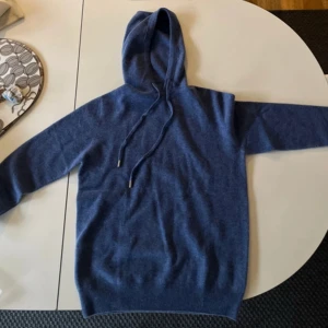 Blå stickad hoodie i merinoull  - Mysig blå stickad hoodie med huva och långa ärmar. Tröjan har ribbade muddar och snörning i halsen för en avslappnad look. Perfekt för dig som gillar stilren och enkel design.