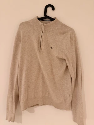 Beige half zip tröja från Gant - Snygg beige tröja från Gant med half zip och ribbad krage. Liten broderad logga på bröstet och långa ärmar. Perfekt för dig som gillar stilrena och klassiska plagg med en chill vibe. Passar folk mellan 165 till 175